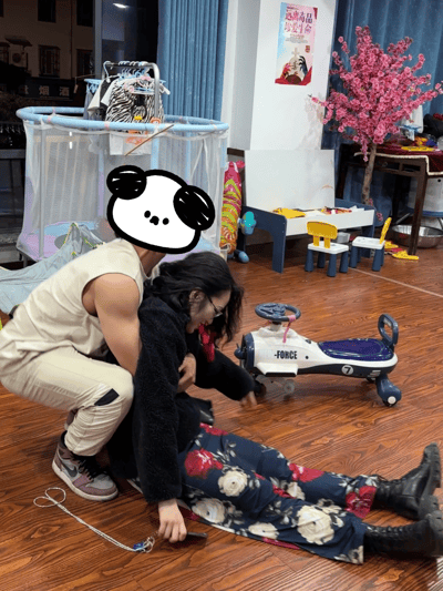 室内场景中，一名肌肉男穿着无袖背心和米色工装裤，正俯身搀扶一位戴眼镜、穿黑色毛绒外套与花裤子的女性。女性身体前倾，伸手似乎想触碰地板上的蓝色'FORCE'玩具车。男子头部被卡通熊猫头贴纸覆盖，表情呆萌。背景可见婴儿围栏、彩色儿童桌椅、粉色假花树及墙上的'远离毒品 珍爱生命'宣传海报，整体氛围温馨又带点搞笑。