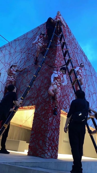 图片展示了一个引人注目的表演艺术现场，在黄昏时分的蓝色天空背景下，一座大型建筑结构的外墙上覆盖着密集的红色网状物。多名穿着白色服装并被红色绑带束缚的表演者正在攀爬这个红色网状结构，他们的面部被模糊处理。有几位身穿黑色服装的工作人员在地面上用长梯子协助他们攀爬，梯子斜靠在建筑上，形成了主要的攀爬路径。整个场景营造出一种紧张而富有戏剧性的氛围，结合了人体艺术、装置艺术和表演元素。