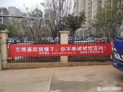 这是一张在城市居民区户外拍摄的照片，画面中铁栅栏上悬挂着一条醒目的红色征兵宣传横幅，横幅上用接地气的网感话术写着“兰博基尼弱爆了，你不来试试坦克吗？”，同时标注了官方征兵网址和“东新乡人民武装部 宣”的字样。背景可见高层居民楼、树木和停放的车辆，画面右下角露出一辆蓝色卡车的局部。这条标语打破了传统征兵宣传的严肃风格，将年轻人熟知的豪华跑车兰博基尼与军队的坦克进行对比，用幽默新潮的方式吸引适龄青年关注征兵，激发他们的参军兴趣，是一种贴合当下年轻人语境的创新宣传方式。