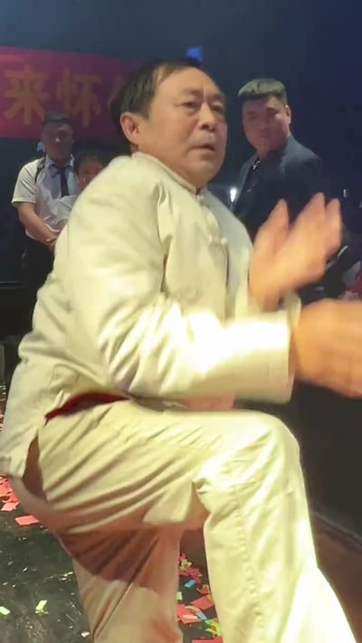 图片中显示马保国身着白色传统武术服装，正在表演其标志性的'闪电五连鞭'动作。他身体微微下蹲，双手做出鞭打的姿势，表情专注。背景中有几位观众在观看，地面上散落着红色的纸屑，后方有一个红色的横幅，上面有部分可见的中文字符。这一场景捕捉了马保国在公开场合展示其武术技巧的瞬间，该动作后来因网络传播而成为流行梗。