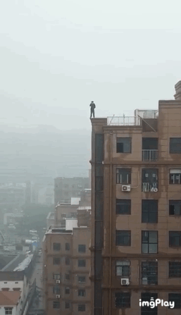 这是一段趣味视觉错位的动态GIF，开篇画面中一名男子站在高层居民楼的楼顶边缘，姿态看起来像是要跳楼，让观看者产生惊险的错觉；随后男子向前跃出，镜头同步移动，揭晓真相——男子只是跳到了紧邻的一栋矮楼楼顶，利用两栋楼的高度差和拍摄角度营造出的视觉错觉，制造出虚惊一场的乌龙效果，整体是充满反差感的搞笑整蛊内容。