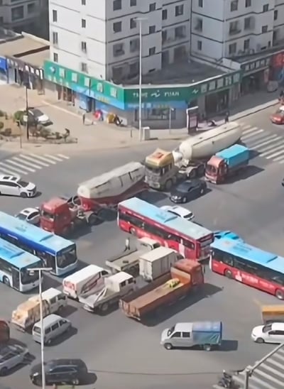 这是一张从高空视角拍摄的城市十字路口交通场景，画面中水泥罐车、公交车、大小货车、私家车等各类车辆在路口互不相让、抢行通过，最终挤成一锅粥，导致路口完全瘫痪，交通彻底堵塞。路边可见少量行人，整个场景十分混乱，直观展现了城市交通中抢行引发的恶劣后果，无谐音梗或双关含义。