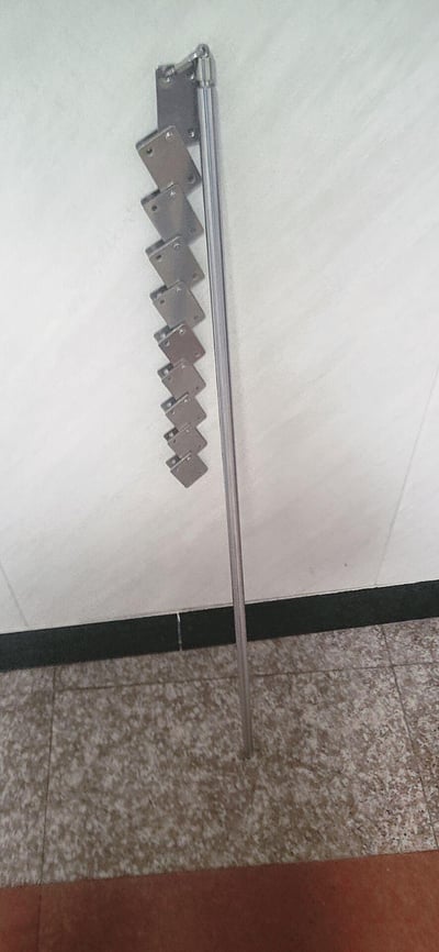 这是一张室内场景实拍照片，展示了一款安装在墙角的折叠式金属辅助扶手。该扶手由多段可折叠的金属支架和一根长金属杆构成，常态下可折叠贴合在白色墙面上，展开后能够为行动不便的人群（如老年人、残障人士）提供行走支撑，适合放置在卫生间、走廊等需要辅助行走的区域。图片下方是带有斑驳花纹的地砖，墙角处设有黑色踢脚线，整体场景简洁实用。
无谐音梗、双关或暗示类内容。