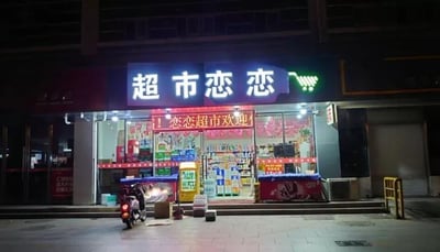 这是一张夜晚拍摄的超市门面照片。超市招牌为蓝底白字的‘超市恋恋’，右侧配有绿色购物车图标。下方红色电子屏滚动显示‘恋恋超市欢迎您’。店内货架摆满各类商品，灯光明亮，门口停放着一辆摩托车，地面散落几个纸箱，整体呈现出社区超市夜间正常营业的日常温馨场景。