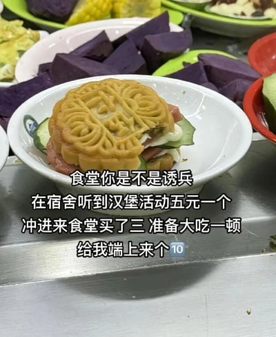 这张图片拍摄的是食堂的一款创意餐品，将月饼作为汉堡的上下层面包，中间夹入火腿片、黄瓜片和白色酱料，放在白色小瓷碗中，外观乍一看类似汉堡。背景里能看到食堂的其他餐食，比如切块的紫薯、其他餐盘里的食物。图片配文是一位学生的吐槽：在宿舍听到食堂有五元一个的汉堡活动，兴冲冲买了三个准备享用，结果拿到的却是月饼做的“汉堡”，用“诱兵”来调侃食堂的营销方式，最后用数字“🔟”来表达自己的无奈与哭笑不得，整体充满校园生活的趣味感，展现了食堂餐品的奇葩创意。