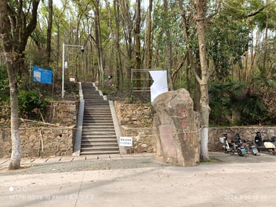 这是2024年3月30日拍摄的武汉喻家山一处登山入口实景：画面中央是通往山林的石阶登山道，被警示带与“喻家山修危枝 行人请勿进入”的提示牌封闭；右侧立着刻有“喻家山”红色字样的景观石，石面还有小字碑文；道旁石墙、周边树木林立，部分树木枝叶稀疏，显示正处于树木维护阶段；画面右下角停放着三辆电动车，左下角标注了拍摄设备为REDMI NOTE 10 PRO，右下角标注拍摄时间为2024.3.30 15:25。
