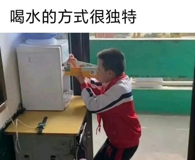 这是一张原始拍摄的校园场景照片，画面主体是一名穿着红白相间校服、系着红领巾的小学生，他身处室内靠近窗户的位置，面前的黄色桌子上摆放着一台白色饮水机。这名小学生没有使用常规的水杯接水，而是用一个长方形的文具收纳盒（外观类似笔袋）承接饮水机的出水，随后将脸凑到收纳盒边缘饮用盒中的水，这个非常规的喝水方式显得十分滑稽有趣。图片上方配有文字“喝水的方式很独特”，精准点出了这个场景的笑点，展现出孩子天马行空、不拘小节的可爱行为，充满了校园生活的趣味性。
