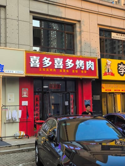 图片展示了一家名为“喜多喜多烤肉”的街边餐馆门面。红色招牌上写有“土耳其烤肉 盖薯条 盖饭 卷饼”等菜品。店门左侧有“科技护肤 全国连锁”的店铺，右侧是黄色招牌的“李”姓店铺。门口挂着几件白色衣物，玻璃门上贴有红色对联“来無代數念”。一名男子站在店门口，前方停着一辆黑色轿车，背景是现代风格的建筑物。