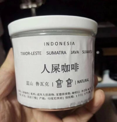 这是一张实拍的恶搞罐装咖啡产品图，罐身标签以白色为底，上方印有英文的印尼相关产区名称“INDONESIA”“TIMOR-LESTE SUMATRA JAVA SUIAWESI”，核心大字为“人屎咖啡”，下方标注了“蓝山 鲁瓦克”、咖啡器具图案以及“NATURAL”字样。标签底部的配料表十分荒诞，写着“成年人排泄物，家猫排泄物，咖啡豆”，同时标注了净含量80克、贮存条件为阴凉干燥、产地印度尼西亚、保质期1个月，生产日期起始为2023年12月09日。该产品明显是模仿正经的猫屎咖啡（鲁瓦克咖啡）制作的整蛊恶搞商品，用离谱的配料和名称制造荒诞搞笑的效果，无相关双关或谐音梗，纯粹以无厘头的设定博人一笑。