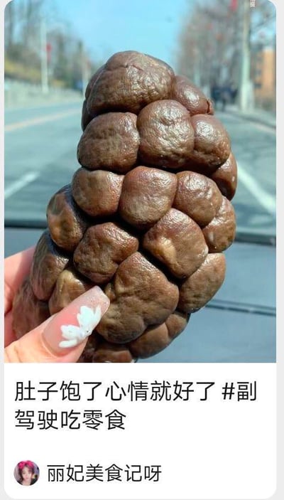 画面中一只做了白色花朵款式美甲的手，在汽车副驾驶位置拿着一个造型十分奇特的深棕色食物，这个食物由多个类似香菇伞盖的凸起紧密排列组成，看起来像是经过腌制的酱菜类零食。下方是抖音的帖子界面，配文写着“肚子饱了心情就好了 #副驾驶吃零食”，底部标注了作者账号“丽妃美食记呀”。整体内容传递出在日常出行时吃零食能带来好心情的轻松氛围，没有双关、谐音梗或暗示内容。