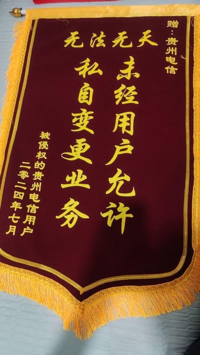 这是一面由被侵权的贵州电信用户制作的讽刺式维权锦旗，锦旗主体为酒红色配金色流苏与边框，用金色字体书写内容，抬头标注“赠：贵州电信”，核心内容以“无法无天”为总评，直指贵州电信“未经用户允许，私自变更业务”的行为，落款为“被侵权的贵州电信用户”与“二零二四年七月”，以此表达用户对运营商违规操作的强烈不满与抗议。