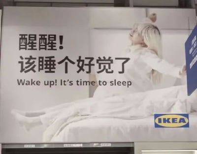IKEA广告海报，画面中一名金发模特身着白色衣物，躺在铺有白色床单的床上伸展身体，表情放松。海报中央有醒目文字：“醒醒! 该睡个好觉了”及英文翻译“Wake up! It's time to sleep”，底部右侧有IKEA蓝色标志。文案通过“醒醒”与“睡觉”的矛盾组合制造幽默效果，旨在推广宜家的睡眠产品，传递舒适睡眠的理念。