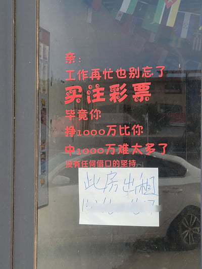 这是一张实地拍摄的竖版照片，主体是一家彩票店的玻璃门。玻璃上用醒目的红色艺术字写着极具调侃意味的彩票宣传语：“亲：工作再忙也别忘了买注彩票 毕竟你挣1000万比你中1000万难太多了 没有任何借口的坚持”，以反讽职场人赚钱不易的方式，吸引路人关注并购买彩票；宣传语下方贴着一张手写的白色纸条，内容为“此房出租”，纸条上的联系电话被做了马赛克处理。玻璃门还反射出店外的白色轿车、建筑等街景，玻璃顶部能看到一些各国国旗的装饰，整体显示出这家彩票店铺目前处于待出租的状态。