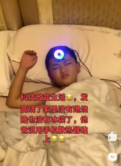 整张图为真人实拍的生活场景，一名小男孩正躺在床上，盖着被子闭眼休息，看起来处于发烧的不适状态，他的额头上没有用常规的退烧贴，反而贴了一个亮着蓝紫色灯光的手机散热器。图上叠加红色文字配笑哭表情说明场景背景：孩子发高烧时家里没有退烧贴和冰袋，孩子的父亲想到了用手机散热器贴在额头上帮助降温，整个内容充满生活化的搞笑脑洞感，右下角还有社交媒体的点赞、评论互动按钮。