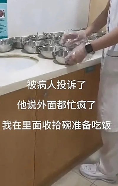 这是一张医院场景的实拍图，画面右侧是身着白色医护工作服、戴透明手套的医护人员，正站在操作台旁整理不锈钢餐碗，操作台上摆放了多只同款不锈钢碗，旁边还设有水池。图上叠加了三行白色配文，是图中医护人员的吐槽内容：自己在后台收拾碗准备吃饭，却被病人投诉，投诉理由是外面就诊区域已经忙疯了，医护却在忙私事，展现出医护工作者工作辛苦却被患者误解的无奈处境。