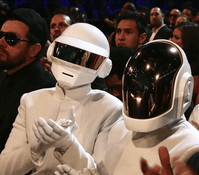 这是一段动态图，展示了法国传奇电子音乐组合蠢朋克（Daft Punk）的两位成员，戴着他们极具标志性的未来感头盔，身着统一的白色礼服，坐在格莱美颁奖礼的观众席中。画面里左侧的成员正抬手鼓掌，周围环绕着其他出席颁奖礼的嘉宾，整体场景正式又带着蠢朋克独有的科幻复古风格，常被用来表达认可、捧场的情绪，图中无谐音梗或双关内容。