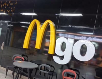 这是一张麦当劳门店的实拍照片，门店墙面上将品牌经典的黄色“M”标志与白色的“go”字样组合，形成“Mgo”的创意标识，下方设置了多组灰黑、红灰配色的圆形桌椅，供顾客休息或等候取餐。该标识设计简洁直观，突出了快捷取餐、外带的服务属性，没有双关或谐音梗内容。