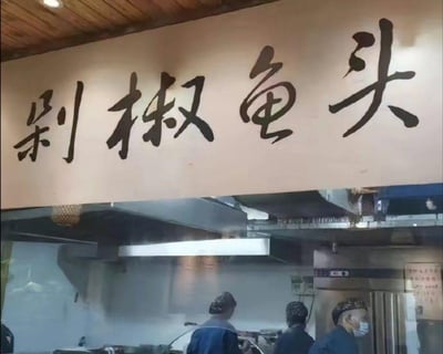 图片展示了一家餐厅的内部场景，上方悬挂着一块招牌，上面用黑色毛笔字书写着“剁椒鱼头”四个大字，字体苍劲有力。招牌下方是厨房区域，几位穿着蓝色制服、戴着发网的工作人员正在忙碌，厨房内有蒸汽升腾，呈现出一派热火朝天的烹饪景象。天花板采用木质扣板设计，整体氛围温馨朴实，突出了中式餐厅的传统风格。