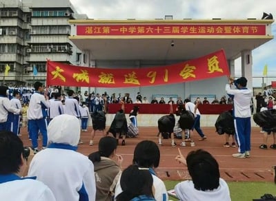 这是湛江第一中学第六十三届学生运动会暨体育节的活动现场，场地上一群学生正在进行创意趣味表演：两名学生举着一条写有“大减就送91会员”的红色横幅，下方多名学生弯腰、套着黑色袋子扮演被“削减”的形象，配合横幅文字制造趣味效果。背景是学校的主席台，上方悬挂着“湛江第一中学第六十三届学生运动会暨体育节”的红色横幅，周围还有其他参与活动的学生和围观的同学，现场氛围欢乐活泼。横幅使用了谐音梗，将“大成就送91会员”中的“成”替换为“减”，结合表演动作，让原本普通的标语变得充满创意和笑点。