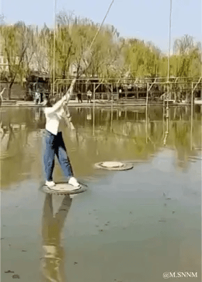 这是一段动态图（GIF），展示了一位身着白色上衣、蓝色牛仔裤的女子，在户外水上区域体验游乐项目的场景。她站在水面上的圆形踏板上，双手紧紧抓住上方悬挂的绳索，努力保持身体平衡，似乎正尝试在这些水上圆盘间移动。背景可见岸边的树木和游乐设施的金属支架，整体环境是充满春日气息的户外水域，画面清晰，呈现出一种惊险又有趣的游玩氛围。图中没有双关、谐音梗或暗示内容。