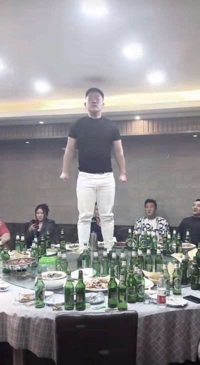 这张图片展现了一位穿黑色T恤和白色裤子的男子，站在餐桌正中央，餐桌上堆满了啤酒瓶，显得极为夸张。桌上有各种下酒菜，但最显眼的是密密麻麻的绿瓶啤酒，似乎现场气氛热烈，显示宴会高潮或某种“哥们义气”的展示时刻。

图片有很明显的“酒桌文化”符号，加上一名男子站桌表演，常作为网络段子调侃饭局上的各种“发酒疯”、“兄弟感情火热”等情况。

没有明显的双关或谐音梗，但有夸张、仪式感强，易被用来讽刺或调侃“喝酒之多”、“饭局荒唐”等场面。