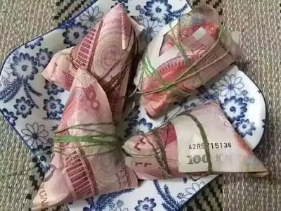 图片中展示了几个用100元人民币纸币折叠成三角形、并用绿色绳子捆绑的包裹物，形似传统粽子，整齐摆放在一个蓝白相间的 floral 花纹陶瓷盘子里，背景是编织纹理的垫子。整体呈现出一种将金钱与节日食品（粽子）结合的创意摄影效果，可能用于节日玩笑或趣味分享，画面构图清晰，色彩对比鲜明。