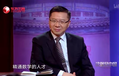 东方卫视《这就是中国》节目截图，一位戴着眼镜、穿着深色西装的男性坐在桌前，面前放着一本打开的书。背景是带有传统中国建筑元素的紫色幕布。屏幕左下角有文字“精通数学的人才”，右下角有节目名称“这就是中国”。