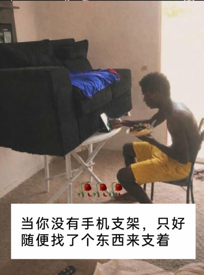 这张图片呈现了一个充满脑洞的生活场景：一名赤裸上身、穿着黄色短裤的黑人男子，坐在房间的椅子上，一边端着装有食物的碗进食，一边操作着被固定在特殊“支架”上的手机。这个“支架”竟是被架在白色折叠小桌上的黑色沙发，男子利用沙发的缝隙来固定手机，以此替代没有手机支架的不便。图片下方配有文字“当你没有手机支架，只好随便找了个东西来支着”，还有三朵凋谢的玫瑰花表情，调侃了这种无奈又搞笑的硬核操作，整体氛围轻松诙谐，展现了生活中突发奇想的趣味瞬间。