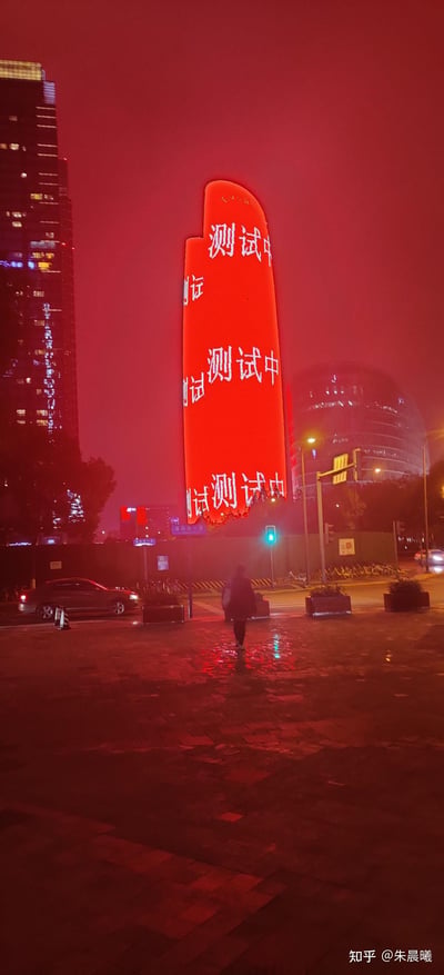 这是一张雨夜的城市夜景实拍图，画面核心是一栋造型独特的弧形建筑，其外立面被大面积红色灯光覆盖，上面重复显示着“测试中”“测试”的白色字样，表明建筑的户外显示屏正在进行测试。地面因降雨变得潮湿，积水反射着红色灯光，增添了夜景的氛围感。前景处有一名行人正走向路口，周围可见其他高楼、亮起的交通信号灯、行驶的车辆以及停放的共享单车，整体环境昏暗，被红色灯光渲染出独特的视觉效果。