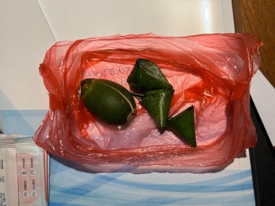 这是一张实拍的日常食物照片，画面中心是套着红色塑料袋的托盘，托盘里放着一颗被纵向切开的青绿色槟榔，旁边搭配着三个用叶子折成三角形状的蒌叶，托盘内侧隐约印有“师范大学”的字样，左下角露出了湿巾包装的一角。槟榔搭配蒌叶是海南、闽南等地区的特色食用方式，通常会搭配石灰等一同咀嚼。