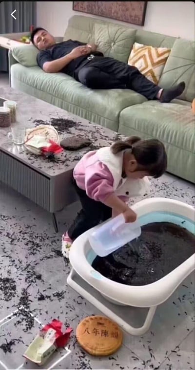 这是一段家庭日常的实拍画面：客厅里，扎着双马尾的小女孩把一块标注“八年陳韻”的茶饼拆开，茶叶撒得满地都是，还将茶叶倒进了一个小浴盆中，正拿着水壶往浴盆里倒水，仿佛在给茶叶“泡澡”。而沙发上的爸爸睡得正香，完全没察觉家里已经被孩子弄得一片狼藉，画面充满了搞笑又无奈的家庭氛围。
