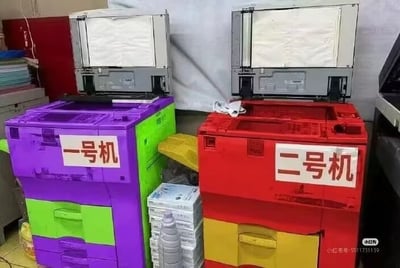 画面里有两台复印机，被夸张地换上了紫绿和红黄撞色的外壳，分别贴着'一号机'和'二号机'的标签。两台机器造型正常但配色极为鲜艳惹眼，完全不同于日常见到的办公设备，看起来非常魔性和喜感，有种'赛博朋克'或'幼儿园风'的感觉。仿佛在暗示两台机器间有某种“竞赛”，让人忍不住联想到选择困难或者PK场景。这个配色和标签组合本身就形成了强烈反差和幽默感，激发了围观者的吐槽欲。