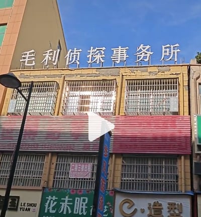 照片展示了一栋临街建筑的上层，黄色墙面搭配铁栅栏窗户，顶部竖立着醒目的白色大字招牌——“毛利侦探事务所”。建筑下层可见“花未眠”、“造型”等商铺招牌，以及“二楼三楼出租 13998083337”的招租信息。画面中央有一个白色播放按钮图标，暗示可能是视频截图。整体场景充满生活气息，招牌名称让人联想到《名侦探柯南》中的经典地点，形成现实与动漫的趣味联动。