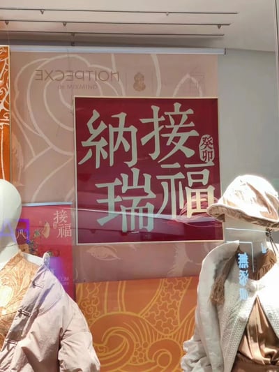 这是一张商场橱窗的照片，展示了春节主题的装饰。画面中有两个穿着浅色冬装的人体模特，背景是带有花纹的装饰板，中央悬挂着一个红色的大型标牌，上面用金色字体写着“纳接瑞福”四个大字，旁边还有“癸卯”字样，表明这是兔年的春节装饰。背景板上还隐约可见品牌名称“EXCEEDING”和“DE MAXMIN”。整体色调温暖，充满节日喜庆氛围。