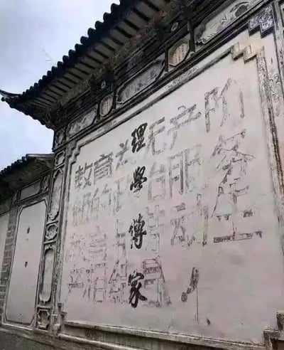 这是一张拍摄中式传统照壁的照片，墙面呈现出明显的文字重叠痕迹：底层是旧时留下的“傳家”等传统字样，上层是后期涂刷的标语类文字，可辨认出“教育为理无产阶”“级的正台服务”“教育与传劳动生”“产相结合”等内容。新旧文字在墙面上叠加，形成了时光流转留下的特殊印记，照壁的中式建筑装饰（如翘角屋檐、雕花边框）也为画面增添了复古氛围。