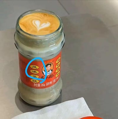 图片中展示了一个玻璃罐，罐身贴有红色标签，标签上印有“风味豆豉”字样及一位人物肖像，通常这类罐子用于盛装咸味的发酵豆制品。然而罐口却一反常态地覆盖着一层咖啡奶泡，奶泡上还精心制作了心形拉花，形成强烈的视觉反差——本应是 savory 风味的调味品容器，却呈现出 sweet 咖啡饮品的顶部特征，营造出幽默有趣的效果。
