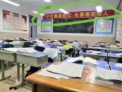 这是一张拍摄于中小学教室的实拍图片，教室内整齐摆放多排课桌椅，桌面散落着书本、试卷、练习册等各类学习资料，教室后方黑板上方悬挂有一条红底黄字的横幅，内容为“宁可累死自己，也要卷死别人”，该横幅被后期添加的绿色圆圈特别标注，黑板两侧还贴有学生学习规范类的宣传海报，整体呈现出浓厚的学习氛围，横幅内容则以夸张的方式调侃了当下学生群体中存在的学业内卷现象。