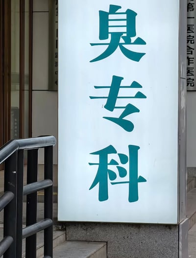 这是一张实拍的医院户外场景照片，画面里的白色竖版科室标识牌原本完整内容是“耳鼻喉专科”，但标识的左侧被黑色扶手栏杆完全遮挡，仅露出右侧的“臭”“专”“科”三个大字，看上去仿佛标识写着“臭专科”，形成了充满笑点的视觉巧合，背景还能看到医院建筑的其他配套标识和台阶设施。
