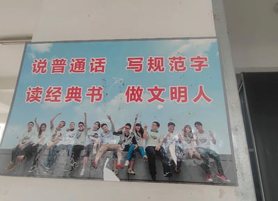 这是一张实拍的公共区域宣传海报照片，海报整体为校园文明宣传主题，上方蓝天背景区域印有红底白边的标语“说普通话 写规范字 读经典书 做文明人”，标语下方是一排穿着同款浅色T恤、姿态活泼的青年学生合影。但海报表面沾染了多处黑、白色的污渍，形态类似鸟类排泄物，恰好落在标语文字上和下方学生形象的头部、身体位置，与“做文明人”的宣传主旨形成极具反差感的荒诞效果，十分滑稽。