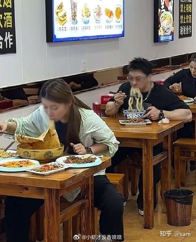 这张实拍图取景于一家西北风味面馆内，最具反差感的是近处女性食客面前摆放的尺寸远超常规的巨型面碗，她正夹起高出碗面很多的面条进食，桌上还摆有多份小菜；她身后的男性食客也正大口嗦着粗面条，手边放有搭配面食的生蒜，墙面挂着餐品菜单灯箱和禁烟禁酒的提示，整体场景充满生活化的喜感，直观展现出西北餐饮分量充足的特点。