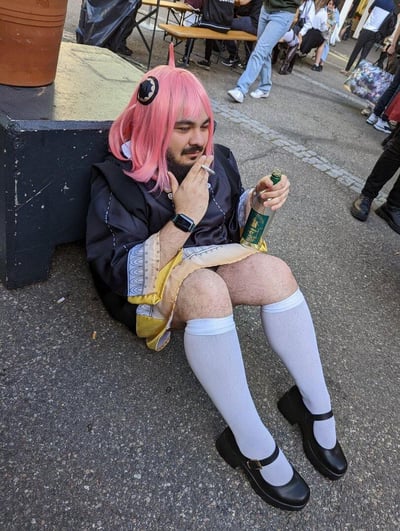 一位留着胡子的男子坐在户外地面上，头戴粉色假发，身穿黑色连衣裙和白色长袜，脚踩黑色皮鞋。左手夹着香烟，右手拿着一瓶绿色标签的酒，表情慵懒，周围有行人经过，背景是街道和一些桌椅。这种强烈的反差萌造型十分吸睛，展现出一种不拘一格的随性气质。