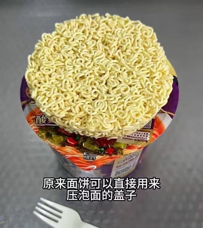 图中展示了一个打开的桶装老坛酸菜泡面，没有使用自带的盖子，而是将干的泡面面饼直接放置在桶口，用来压住桶内的料包，下方配有文字注解“原来面饼可以直接用来压泡面的盖子”，分享了一个实用又新奇的泡面小技巧，无双关、谐音梗或暗示内容。