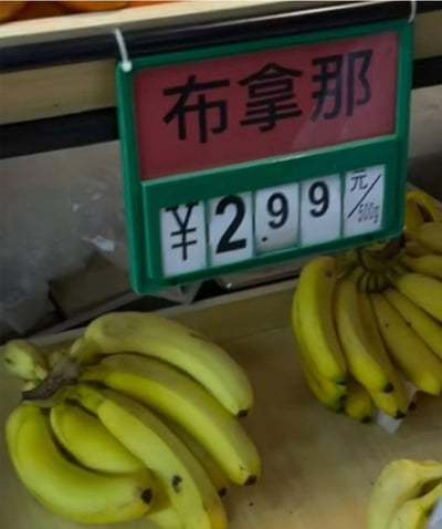 这是一张超市水果售卖区的实拍静态图，画面里的货架上摆放着几串表皮鲜黄的香蕉，看起来新鲜饱满。在香蕉上方的绿色边框标价牌中，红色区域写着商品名“布拿那”，下方白色区域标注着价格为¥2.99元/500g。这里运用了谐音梗，“布拿那”是英文单词“banana”的音译，用这种有趣的方式给香蕉命名，让普通的商品标价变得诙谐有趣，增添了生活中的小趣味。