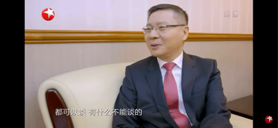 这是东方卫视的一段正式访谈画面，学者张维为身着深色西装搭配红色领带，佩戴眼镜，坐在浅色沙发上，面带温和的微笑，侧身进行交流。画面下方配有字幕“都可以谈 有什么不能谈的”，传递出开放包容的交流态度，场景为装修正式的访谈室内，背景有木质腰线与浅色墙面。