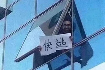 这是一张原始拍摄的静态单图，画面里一名留着胡须的男子身处现代化玻璃幕墙写字楼的打开的窗内，他从窗户探出身子，双手举着一张手写简体中文“快逃”的白纸，脸上带着无奈的笑容，背景是蓝色的玻璃幕墙与晴朗的蓝天。这张图被用作职场相关的梗图，以夸张的方式传递出职场人想要逃离工作压力、摆脱职场困境的情绪，能够引发职场群体的情绪共鸣，没有双关、谐音梗等特殊内容。