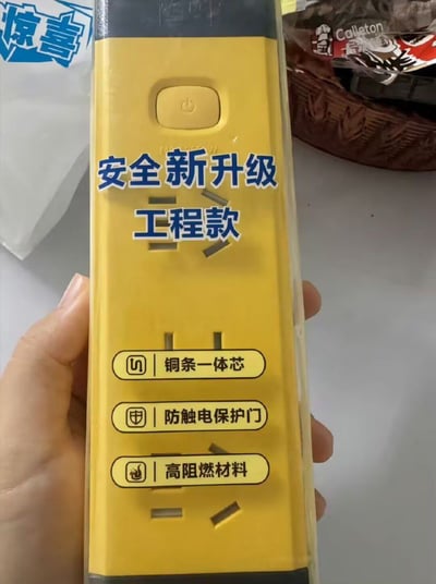 一只手拿着一款黄色的工程款插排，插排包装上清晰标注了「安全新升级 工程款」的字样，还列出了「铜条一体芯」「防触电保护门」「高阻燃材料」三项安全相关的产品特性。画面左上角有一个蓝色的「惊喜」贴纸，背景中能看到一个装有Calleton品牌食品的篮子，整体是对这款工程用插线板的实拍展示，突出其安全升级的特点。