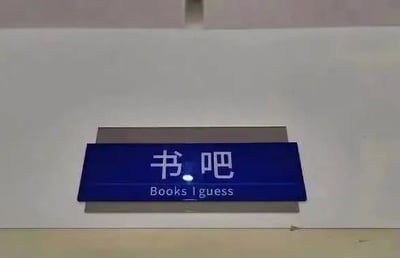 一张拍摄于室内的招牌照片，画面中央是一块蓝色长方形标牌，固定在白色墙面上。标牌上方用白色简体中文书写“书吧”二字，下方配有英文翻译“Books I guess”。英文翻译中的“I guess”（我猜）带有随意、不确定的语气，与正式的中文招牌形成幽默反差，显得轻松有趣。
