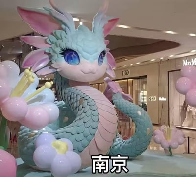 这是2025农历蛇年期间，南京某高端商场内设置的春节主题美陈。主体是一只造型软萌的Q版青粉色蛇形雕塑，有着大大的星空蓝眼睛和飘逸的粉内耳状角，蛇身覆盖着青绿色鳞片，腹部为淡粉色，身体盘绕着，周围搭配粉蓝渐变的花朵装饰。背景可见FENDI、MaxMara等奢侈品牌的门店，整体风格可爱治愈，用来烘托蛇年新年的喜庆氛围，吸引顾客打卡。