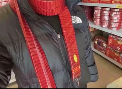 图片中显示一个人穿着黑色North Face夹克，脖子上围着用红色鞭炮串成的"围巾"，背景是摆满红色包装商品的货架。这张图通过将鞭炮创意性地用作围巾，并结合文字"过年了，兄弟借个🔥，我围巾有线头要烧一下"形成幽默效果，暗示想点燃鞭炮而非真的烧线头。