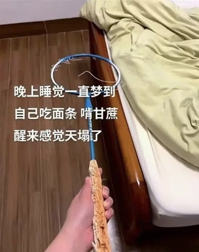 图片展示了一只手握着一个羽毛球拍，拍柄部分有明显被啃咬过的痕迹，呈现出不规则的破损边缘。图片左侧配有文字：“晚上睡觉一直梦到自己吃面条 啃甘蔗 醒来感觉天塌了”，生动描述了因做梦吃东西而在现实中误咬坏物品的搞笑场景。整体画面以卧室为背景，木地板和床边的绿色被子增加了生活气息，营造出一种真实又荒诞的幽默效果。